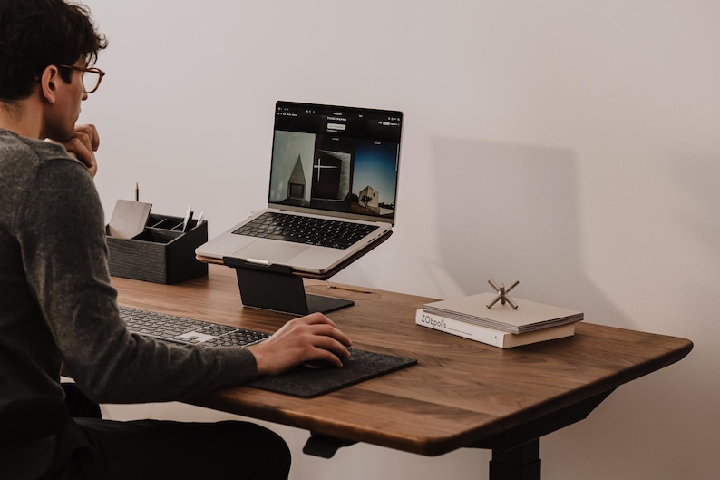 May include: A dark wood desk featuring a silver laptop on a black metal stand, a wireless keyboard, a dark gray mouse pad with a mouse, a black desk organizer, and a stack of two white books with the text ZOEpolis and a metal jack ornament.