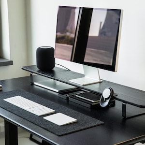 May include: A black desk with a black monitor stand, a black speaker, a white keyboard, a white mouse, and a black mousepad. The desk is made of wood and has a black metal frame.