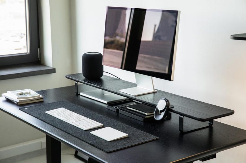 May include: A black desk with a black monitor stand, a black speaker, a white keyboard, a white mouse, and a black mousepad. The desk is made of wood and has a black metal frame.