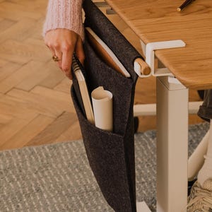 May include: A dark grey felt desk organiser with multiple pockets, attached to a wooden desk. The organiser holds notebooks and rolled paper. The desk has a white metal frame and is near a rug and wooden floor.