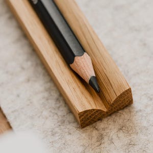 May include: A black pencil rests in a wooden pencil holder. The holder is made of light brown wood and has a slightly curved shape.