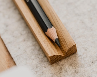 May include: A black pencil rests in a wooden pencil holder. The holder is made of light brown wood and has a slightly curved shape.