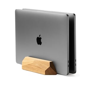 May include: A wooden laptop stand with a geometric design, holding two closed silver laptops. The stand is made of light brown wood and has a natural finish.