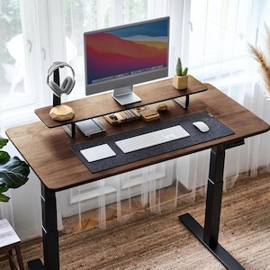 May include: A dark brown wooden standing desk with a black metal frame. The desk has a black mousepad, a white keyboard, and a small potted plant on a wooden shelf. A computer monitor is on the shelf, and a pair of headphones are on a stand.