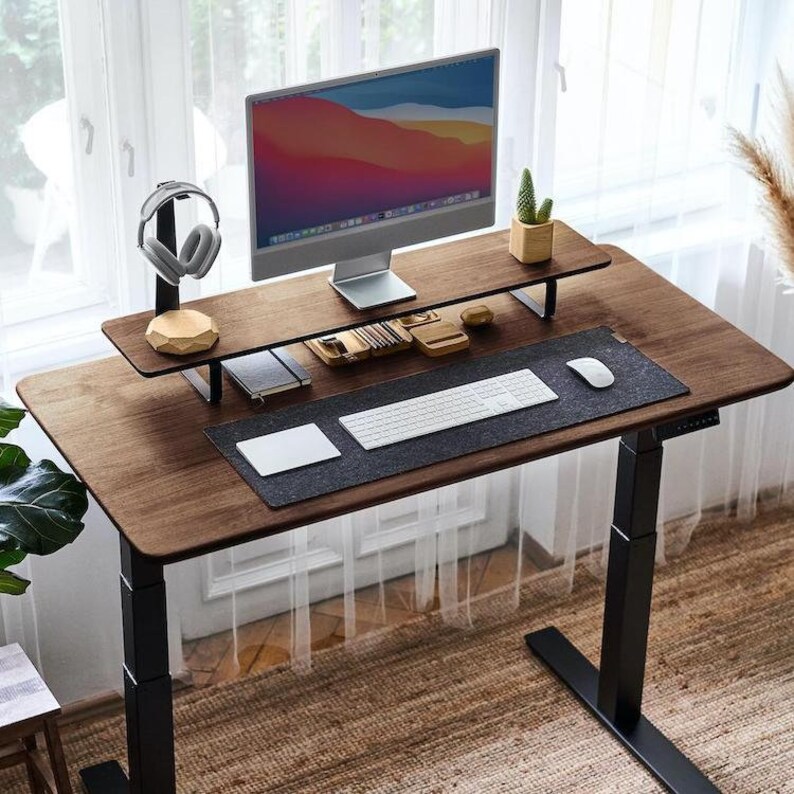 May include: A dark brown wooden standing desk with a black metal frame. The desk has a black mousepad, a white keyboard, and a small potted plant on a wooden shelf. A computer monitor is on the shelf, and a pair of headphones are on a stand.