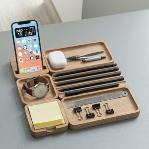 K&ouml;nnte beinhalten: Ein h&ouml;lzerner Schreibtisch-Organizer mit mehreren F&auml;chern zur Aufbewahrung verschiedener Gegenst&auml;nde, darunter ein Telefon, eine Uhr, Stifte, Bleistifte, ein Lineal und B&uuml;roklammern. Der Organizer ist aus hellbraunem Holz gefertigt und hat eine nat&uuml;rliche Oberfl&auml;che.