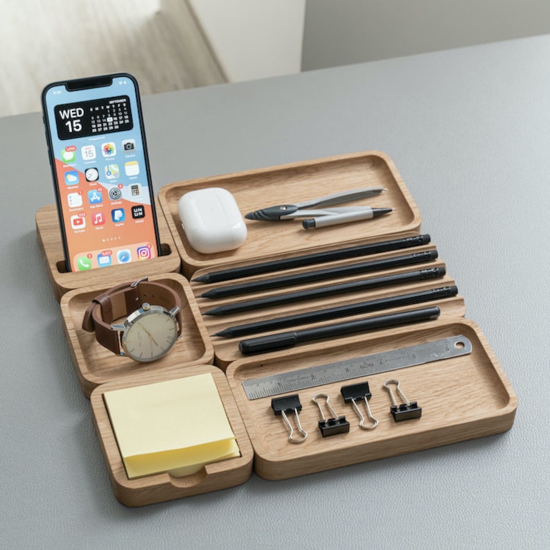 K&ouml;nnte beinhalten: Ein h&ouml;lzerner Schreibtisch-Organizer mit mehreren F&auml;chern zur Aufbewahrung verschiedener Gegenst&auml;nde, darunter ein Telefon, eine Uhr, Stifte, Bleistifte, ein Lineal und B&uuml;roklammern. Der Organizer ist aus hellbraunem Holz gefertigt und hat eine nat&uuml;rliche Oberfl&auml;che.