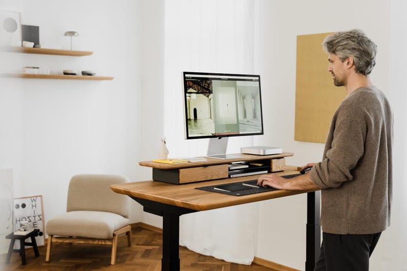 May include: A modern, height-adjustable wooden desk with a computer monitor, keyboard, and mouse. The desk is in a bright, minimalist office setting. A person is standing at the desk, using the computer. The desk has a built-in drawer and a shelf for accessories.