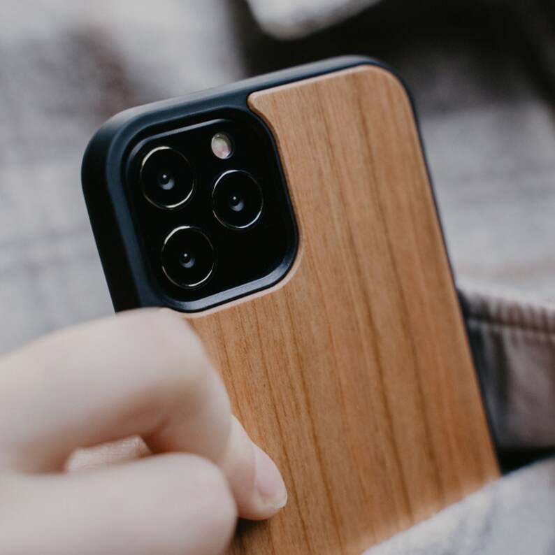 May include: A wooden phone case with a black trim and a camera lens cutout. The case has a natural wood grain pattern.
