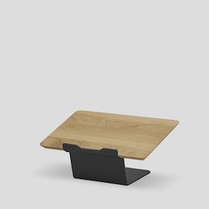 May include: A laptop stand featuring a light-toned wooden platform with a visible grain pattern, supported by a matte black metal base. The platform is angled upward, providing a stable surface for a laptop or tablet device in a workspace.