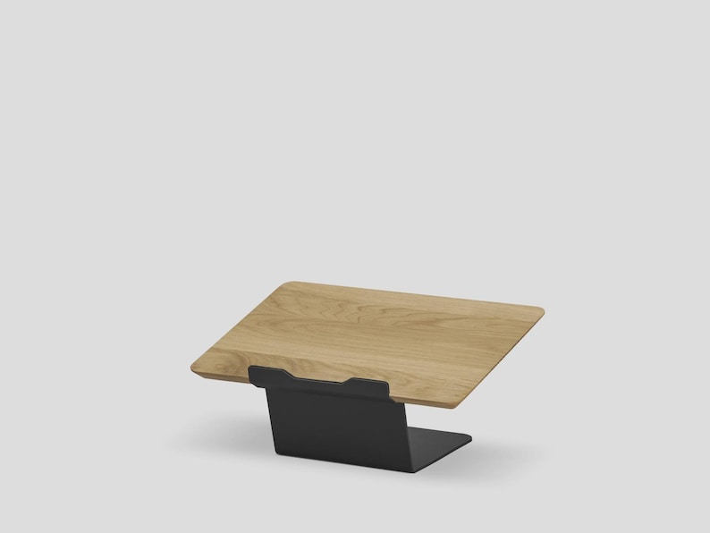 May include: A laptop stand featuring a light-toned wooden platform with a visible grain pattern, supported by a matte black metal base. The platform is angled upward, providing a stable surface for a laptop or tablet device in a workspace.