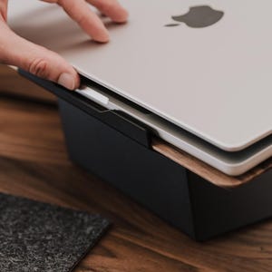 May include: A silver laptop with a black Apple logo on the lid is positioned on a dark wooden and black metal laptop stand. The stand features a flat black base and a slanted wooden platform that supports the laptop at an elevated angle.