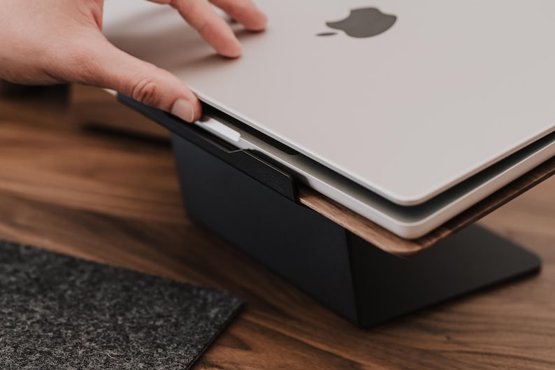 May include: A silver laptop with a black Apple logo on the lid is positioned on a dark wooden and black metal laptop stand. The stand features a flat black base and a slanted wooden platform that supports the laptop at an elevated angle.