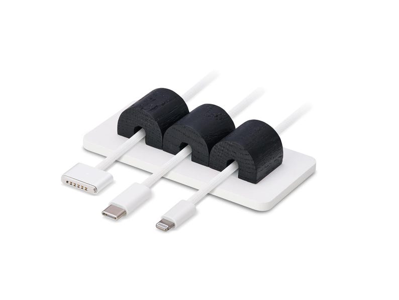 May include: A white cable tidy with three black wooden cable holders. Three white cables are inserted through the holders. The tidy has a rectangular base and is designed to manage and organise cables.