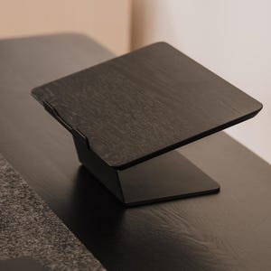May include: A black laptop stand featuring a flat, angled platform with a visible wood grain texture and a solid base. The stand includes a small front lip to secure a device in place, providing an elevated surface for a laptop or tablet.