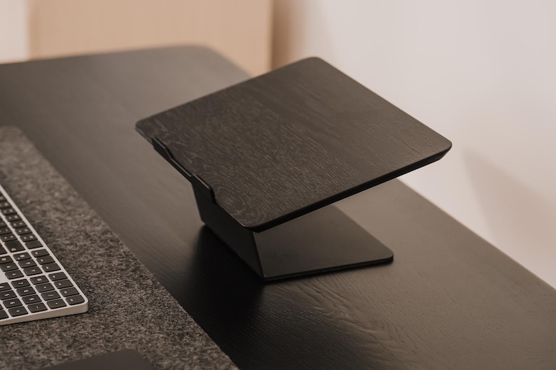 May include: A black laptop stand featuring a flat, angled platform with a visible wood grain texture and a solid base. The stand includes a small front lip to secure a device in place, providing an elevated surface for a laptop or tablet.