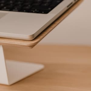 May include: A laptop stand featuring a white metal base and a light-toned wood grain platform. The stand supports a silver laptop with a black keyboard, providing an elevated surface for the device while maintaining a clean and simple structural design.
