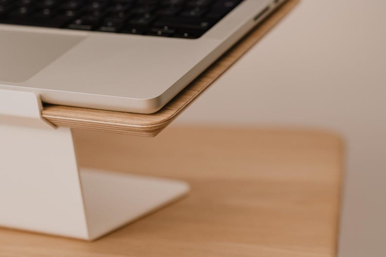 May include: A laptop stand featuring a white metal base and a light-toned wood grain platform. The stand supports a silver laptop with a black keyboard, providing an elevated surface for the device while maintaining a clean and simple structural design.