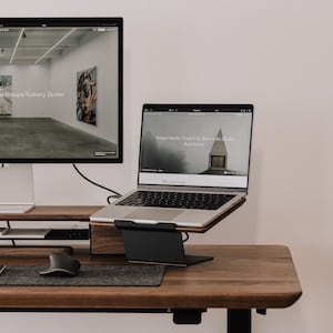 May include: A dark wood desk setup featuring a large computer monitor on a wooden riser, a silver laptop on a black metal stand, a grey felt desk mat, a wireless keyboard, and a black ergonomic mouse. The screens display art gallery websites.