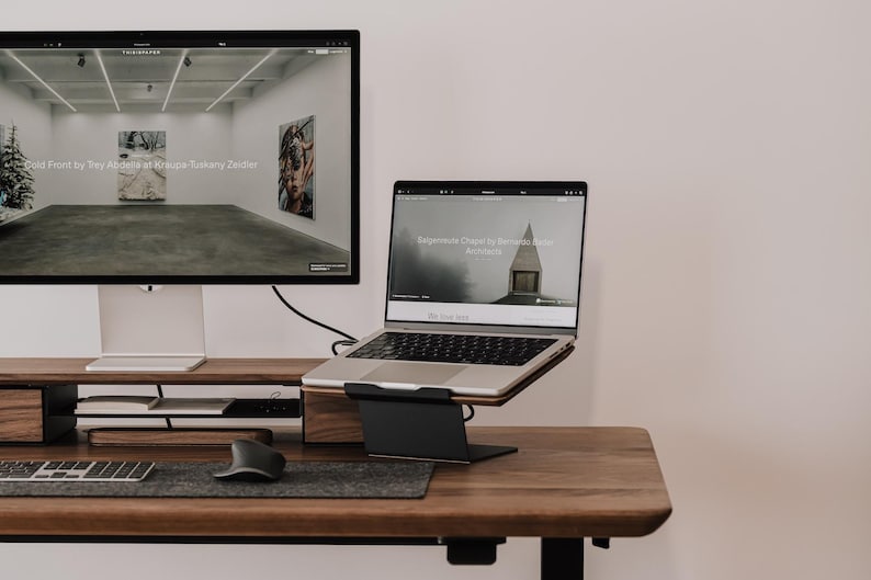 May include: A dark wood desk setup featuring a large computer monitor on a wooden riser, a silver laptop on a black metal stand, a grey felt desk mat, a wireless keyboard, and a black ergonomic mouse. The screens display art gallery websites.