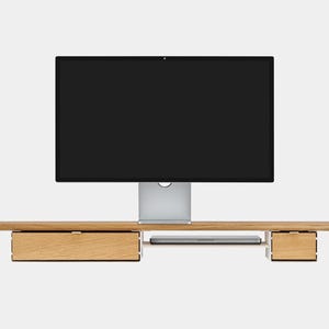 May include: A wooden monitor stand with two drawers and a slot for a keyboard. A large black computer monitor is centered above the stand. The stand is made of light-colored wood.