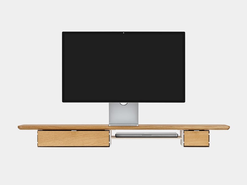 May include: A wooden monitor stand with two drawers and a slot for a keyboard. A large black computer monitor is centered above the stand. The stand is made of light-colored wood.