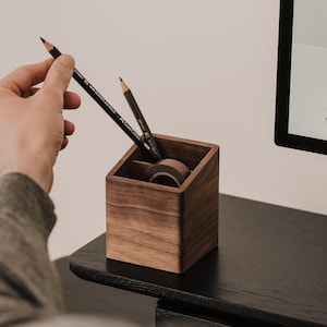 Può includere: Un portamatite quadrato in legno con bordo superiore inclinato, realizzato in legno marrone scuro con venature visibili. Il supporto contiene due matite nere e un rotolo di nastro adesivo, offrendo una soluzione funzionale per riporre articoli da scrivania.