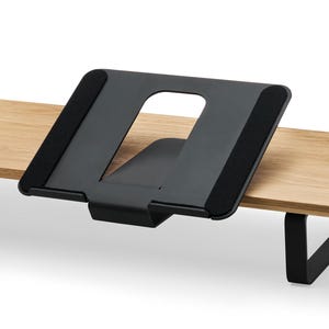 May include: A black metal desk stand with a cut-out center, designed to hold a tablet or book. The stand is mounted on a light-colored wooden surface. The stand has black, padded contact points.