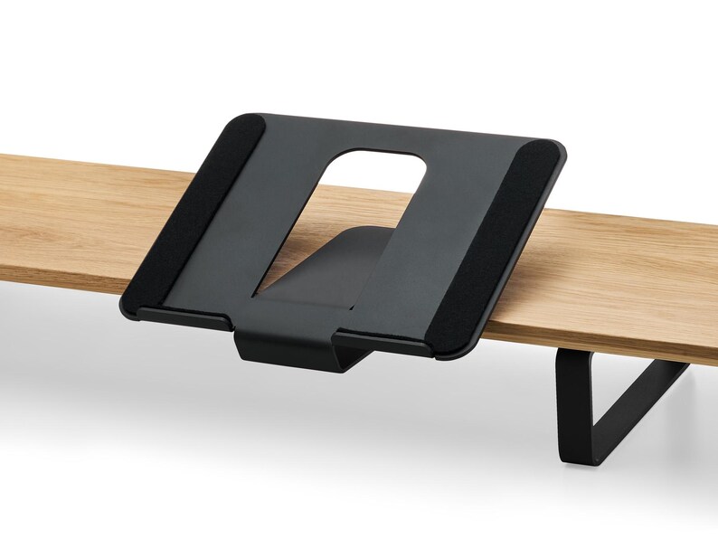 May include: A black metal desk stand with a cut-out center, designed to hold a tablet or book. The stand is mounted on a light-colored wooden surface. The stand has black, padded contact points.