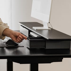 May include: A black standing desk with a monitor, keyboard, and mouse. The desk has a drawer that is being opened. The desk is made of dark wood and has a modern design. The monitor is white.