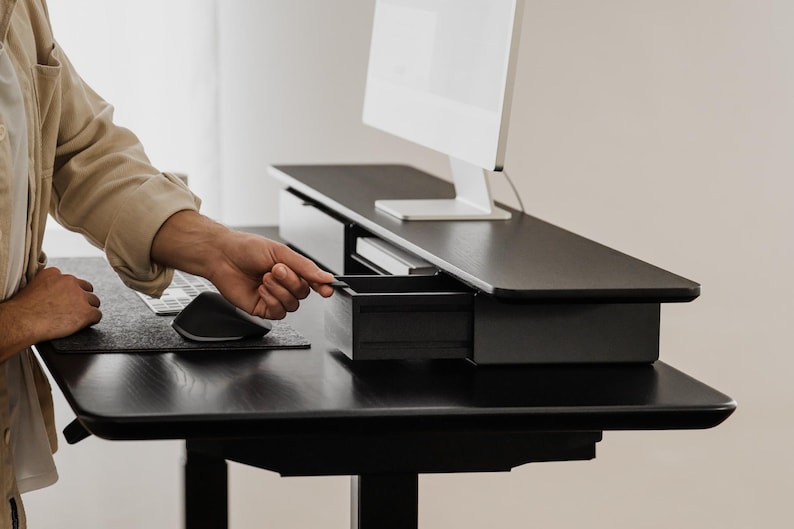 May include: A black standing desk with a monitor, keyboard, and mouse. The desk has a drawer that is being opened. The desk is made of dark wood and has a modern design. The monitor is white.
