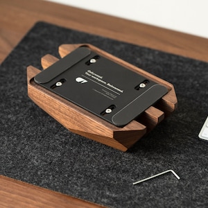 May include: A black metal plate with the text "Oakwood Your workspace, Reinvented" attached to a wooden stand. The stand is made of dark brown wood and has a geometric shape. A small silver Allen wrench is visible on the surface.