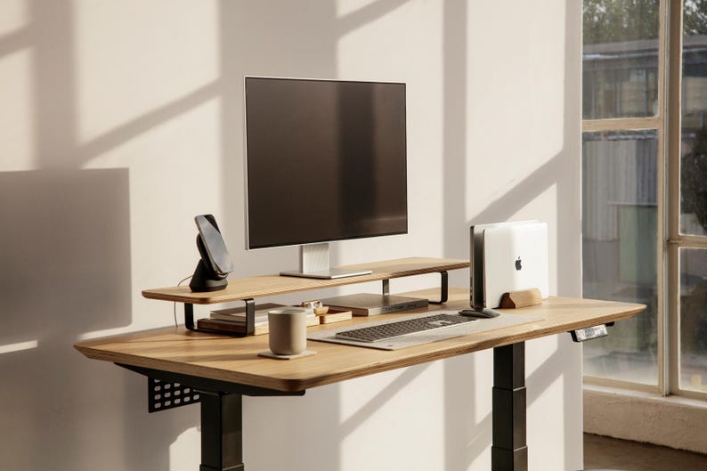 May include: A wooden standing desk with a black metal frame. The desk has a monitor stand with a black phone charger and a white mug on the desk. A laptop is on the desk with a keyboard and mouse.