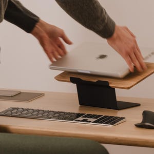 May include: A silver laptop is being placed onto a wooden laptop stand with a black metal base. The setup includes a wireless keyboard and a computer mouse on a wooden desk surface, creating a functional workspace for a portable computer.