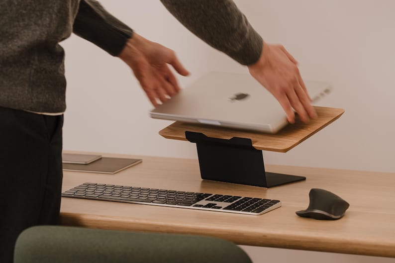 May include: A silver laptop is being placed onto a wooden laptop stand with a black metal base. The setup includes a wireless keyboard and a computer mouse on a wooden desk surface, creating a functional workspace for a portable computer.