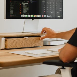 Puede incluir: Un escritorio blanco con un cajón de madera y una superficie de madera clara. Un monitor de computadora está encima del cajón. Una persona está sentada en el escritorio y alcanza el cajón.