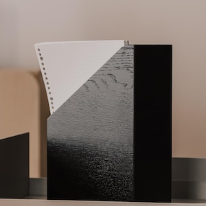 Peut inclure: Un porte-documents en bois noir avec un bord supérieur diagonal, présentant une texture de grain de bois visible. Une feuille de papier blanc avec un bord perforé et un motif quadrillé est insérée dans le support, dépassant du coin supérieur.