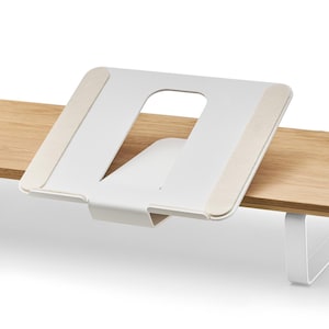 May include: A white metal laptop stand with a cut-out design, mounted on a light-colored wooden surface. The stand has beige pads for grip and a minimalist aesthetic. The stand is designed to elevate a laptop.