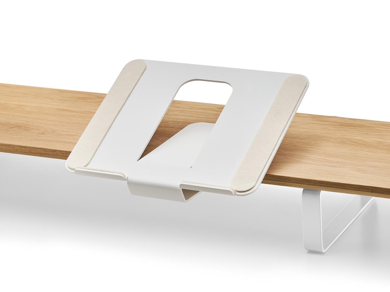 May include: A white metal laptop stand with a cut-out design, mounted on a light-colored wooden surface. The stand has beige pads for grip and a minimalist aesthetic. The stand is designed to elevate a laptop.