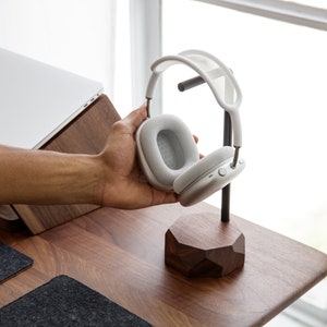 May include: White wireless headphones with a grey earcup on a wooden stand. The stand has a geometric shape.
