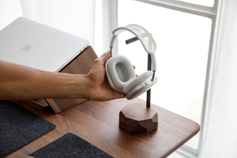 May include: White wireless headphones with a grey earcup on a wooden stand. The stand has a geometric shape.