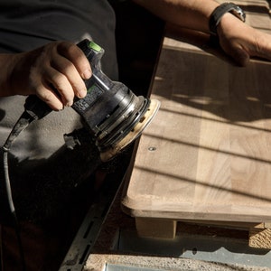 May include: A person sanding a piece of wood with a black and green electric sander.
