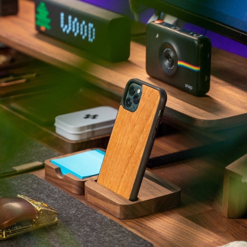 K&ouml;nnte beinhalten: Eine Holz Handyh&uuml;lle f&uuml;r ein iPhone mit einem Ausschnitt f&uuml;r die Kamera. Die H&uuml;lle steht in einem Holzst&auml;nder auf einem Schreibtisch mit einem blauen Haftnotiz und einer kleinen Schachtel.