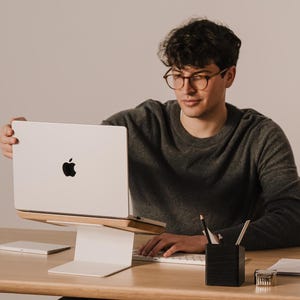 May include: A silver laptop with a black Apple logo on the lid, positioned on a white metal and light wood laptop stand. The stand features a flat base and an angled platform that elevates the device for ergonomic viewing on a wooden desk.