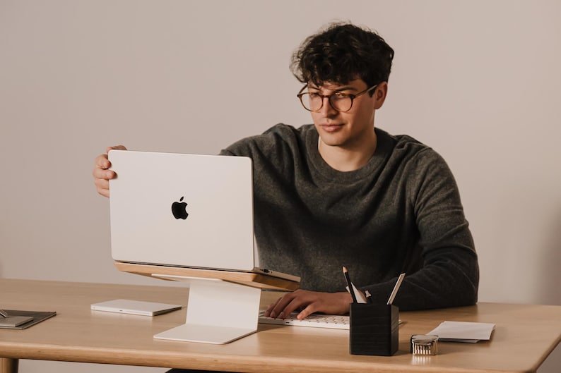 May include: A silver laptop with a black Apple logo on the lid, positioned on a white metal and light wood laptop stand. The stand features a flat base and an angled platform that elevates the device for ergonomic viewing on a wooden desk.