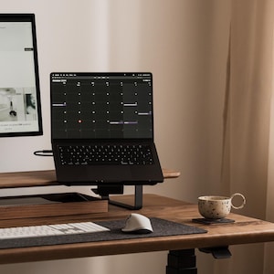 May include: A modern home office setup featuring a black laptop displaying a calendar, a large monitor, and a white keyboard. A wooden desk holds a tray, a mouse, and a patterned mug. The room has a neutral color palette.