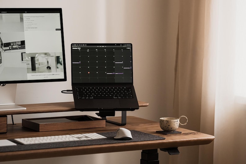 May include: A modern home office setup featuring a black laptop displaying a calendar, a large monitor, and a white keyboard. A wooden desk holds a tray, a mouse, and a patterned mug. The room has a neutral color palette.