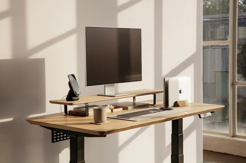 May include: A standing desk with a wooden top and black metal legs. The desk has a monitor stand with a monitor on top, a wireless charging pad, a laptop stand, a keyboard, and a mug. The desk is in front of a window with a view of a building.