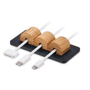 May include: Cable tidy featuring three wooden cable holders on a black base. White cables with various connectors are inserted through the wooden holders. The organiser is designed to manage and organise cables.