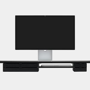 May include: A black computer monitor sits atop a black desk with two drawers on each side. The monitor has a silver stand. A laptop is visible on the desk between the drawers. The desk is mounted on a white wall.